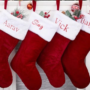 May include: Four red Christmas stockings with white faux fur cuffs are displayed. Each stocking is personalized with a name embroidered in red: Aarav, Sing, Vick, and Baljit. The stockings are hanging from a white mantelpiece, decorated with festive ornaments.