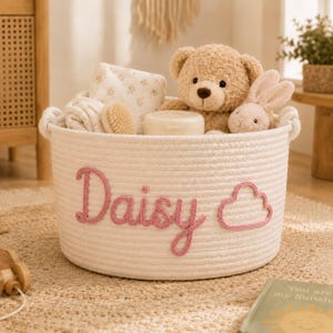 Op de afbeelding: Een ronde opbergmand van wit gevlochten touw met de naam Daisy en een wolkvorm geborduurd in roze garen op de voorzijde. De mand is gevuld met een beige teddybeer, een lichtroze pluche konijn, een witte doek met patroon en een kleine container.