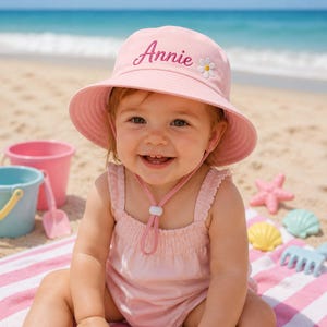 Può includere: Un cappello da pescatore rosa chiaro con il nome Annie ricamato in corsivo rosa scuro e un piccolo fiore di margherita bianco con il centro giallo. Il cappello include un cinturino sottogola rosa abbinato con una chiusura bianca regolabile.