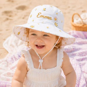 May include: A white bucket hat featuring a small yellow floral pattern and the embroidered name Emma in gold script, accompanied by a single embroidered daisy. The hat includes a white chin strap with an adjustable toggle for a secure fit.