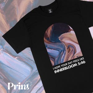 Può includere: Maglietta nera con una grafica astratta di vernice blu, marrone e viola vorticosa a forma di arco. Il testo "I hope your day feels like INNERBLOOM 5:46" è stampato in bianco sotto la grafica.
