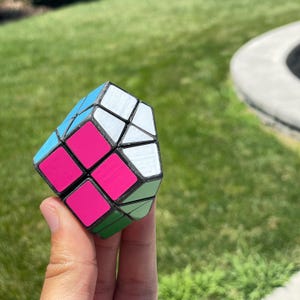 Puede incluir: Un cubo de rompecabezas multicolor con paneles rosas, azules, blancos y verdes. El cubo se sostiene en una mano sobre un fondo verde. El diseño del cubo es geométrico y angular.