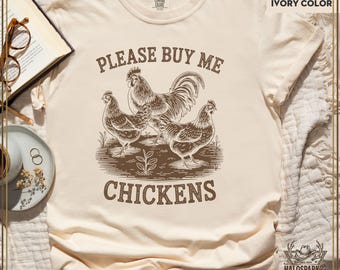 May include: Ivory-colored t-shirt with a graphic of a rooster and two hens, with the text "PLEASE BUY ME CHICKENS" in brown. The shirt is made of a soft material and is a Comfort Colors brand.