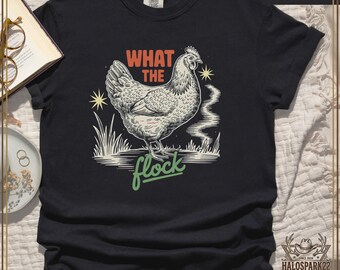 May include: Black t-shirt with a detailed illustration of a chicken, accompanied by the text "WHAT THE flock" in orange and green. The design includes starbursts and stylized smoke. The shirt is a Comfort Colors brand.