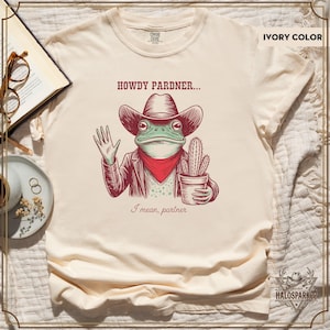 May include: Ivory t-shirt with a cartoon frog in a cowboy hat, red bandana, and jacket, holding a potted cactus. Text reads "HOWDY PARDNER... I mean, partner."