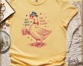 Puede incluir: Camiseta de color mantequilla con una ilustración de un ganso. El ganso lleva gafas de sol, sombrero y pañuelo, sosteniendo una jarra de cerveza. El texto dice "Red, White and Booze" y "This Goose is Loose."