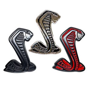 May include: Three cobra emblems in black, silver, and red. Each emblem features a detailed cobra design with a raised hood and a curved body. The emblems are glossy and appear to be made of metal or a similar material.