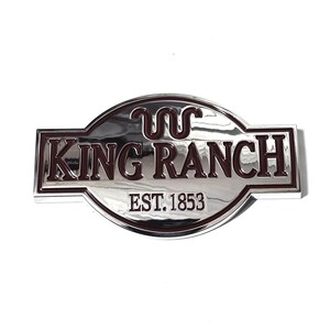 May include: A silver-coloured metal emblem with the words "KING RANCH" in dark red lettering. The emblem has a curved top and bottom with the text "EST. 1853" below the main text. The emblem has a shiny, reflective surface.