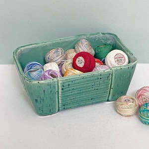 May include: A teal ceramic container holding various colourful thread spools. Several spools are outside the container. The thread colours include red, white, green, blue, and pink. The container is rectangular with a textured surface.