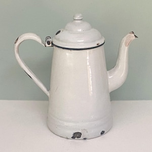 May include: White enamel coffee pot with a lid and a curved handle. The pot has a spout and a black trim around the lid. The enamel shows signs of wear, with some areas of discoloration and chipping, giving it a vintage look.