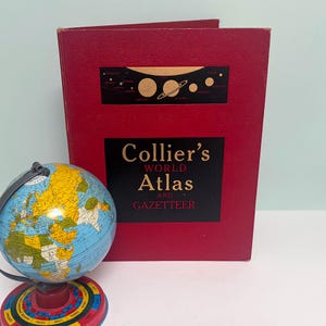 May include: A red hardback book titled Collier's World Atlas and Gazetteer with a gold solar system graphic on the cover, positioned behind a small, colourful desktop globe featuring a circular base with zodiac signs printed around the edge.