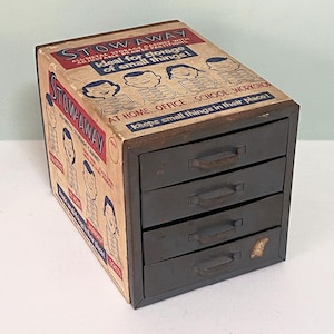 May include: Vintage metal Stowaway storage cabinet with four drawers. The cabinet has a cardboard exterior with the words "Stowaway" and "ideal for storage of small things!" printed on it. The drawers are dark gray.