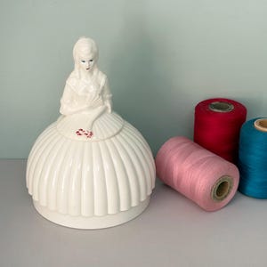 May include: A white ceramic figurine of a person in a wide, pleated skirt, holding a fan and a small red floral detail. Beside the figurine are three spools of thread in solid red, light pink, and teal colours, arranged on a flat surface.
