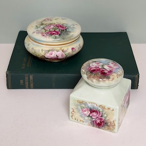 Pode incluir: Duas caixas decorativas de porcelana com desenhos florais. Uma é redonda com tampa, a outra retangular com tampa. Ambas apresentam rosas cor-de-rosa e folhas verdes sobre um fundo creme. As caixas estão sobre um livro verde escuro.