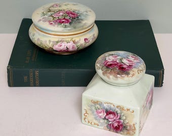 Peut inclure: Deux boîtes décoratives en porcelaine avec des motifs floraux. L'une est ronde avec un couvercle, l'autre est rectangulaire avec un couvercle. Les deux présentent des roses roses et des feuilles vertes sur fond crème. Les boîtes sont sur un livre vert foncé.