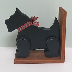 Peut inclure: Un serre-livres en bois noir en forme de chien terrier écossais. La figurine du chien présente un ruban en tissu à carreaux rouges et noirs noué en nœud autour du cou. La base et le panneau de support vertical sont en bois brun naturel.