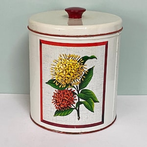 May include: A vintage white metal canister with a red lid and trim. The canister has a rectangular panel with a floral design of yellow and red flowers with green leaves. The panel's background has a crosshatch pattern.