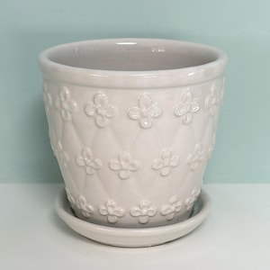 May include: A glossy, light gray ceramic plant pot with a matching saucer. The exterior features a raised, embossed diamond pattern with small, four-petaled flower motifs centered within each diamond shape across the entire surface.
