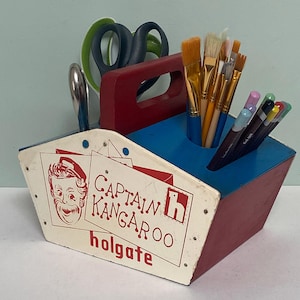 May include: A vintage wooden Captain Kangaroo Holgate toolbox, painted red, white, and blue, holding art supplies. The toolbox features a red handle, and the side displays the Captain Kangaroo logo and text. The toolbox contains paintbrushes, pens, and scissors.