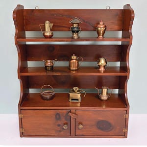 Peut inclure: Étagère murale en bois foncé avec deux étagères et une base d'armoire. Les étagères présentent des miniatures de cuisine en cuivre et en laiton, dont des théières et une lanterne. L'armoire a deux portes avec des boutons en laiton.