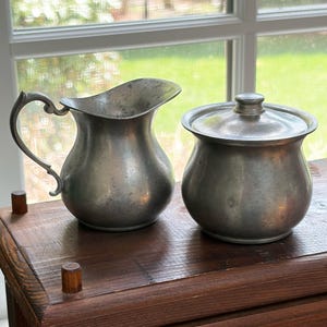 May include: A vintage pewter pitcher with a curved handle and a matching lidded sugar bowl. The antique pieces are displayed on a wooden surface in front of a window, showcasing their classic design and aged patina.