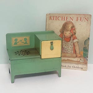 Könnte beinhalten: Ein grüner Spielzeugherd im Vintage-Stil mit gelbem Schrank und dekorativer Silhouette. Daneben ein Buch mit dem Titel "KITCHEN FUN" mit einer Illustration eines kochenden Mädchens. Ein charmantes Stück für Sammler.