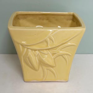 May include: A square-shaped ceramic planter with a glossy pale yellow finish. The exterior features a raised, molded relief pattern of stylized leaves and stems that wrap around the front face of the vessel, which has a wide, open top rim.