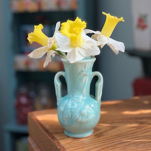 Puede incluir: Un pequeño jarrón de cerámica azul claro con dos asas, que contiene varios narcisos. Los narcisos tienen pétalos blancos y centros amarillos. El jarrón tiene un diseño floral y está sobre una superficie de madera.