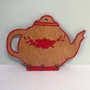 Puede incluir: Llavero de madera con forma de tetera, con acabado marrón claro y detalles pintados en rojo. Presenta un diseño floral rojo y tres ganchos metálicos para llaves. Un artículo decorativo para el hogar.