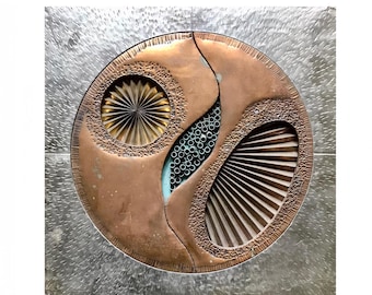 May include: A copper-toned, abstract wall sculpture with a circular design. The sculpture features a textured, hammered metal background. The central design includes a fan-like element, a circular element, and a section with small, circular accents.