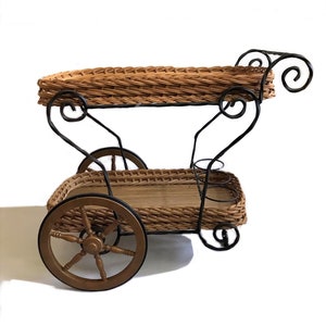 May include: A two-tiered wicker serving cart with black metal frame and wheels. The cart has a woven wicker top and bottom tray with a black metal handle. The wheels are wooden with a black rim.