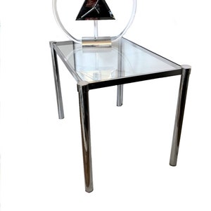 May include: A chrome and glass end table with a triangular clock featuring a black face and a clear acrylic circle around it.