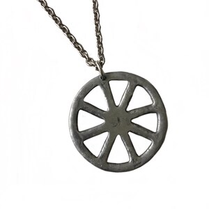 May include: A silver necklace with a circular pendant featuring a wheel design with eight spokes. The pendant has a textured surface and a small inscription on the back.