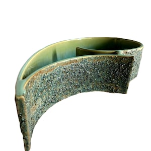 May include: A crescent-shaped ceramic bowl featuring a smooth, light green interior glaze and a contrasting exterior surface with a rough, dark, and speckled textured finish. The vessel has a curved, elongated form with a central divider.