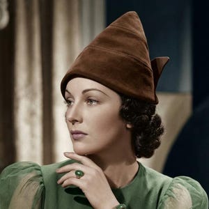 May include: A brown suede pointed hat with a small bow detail at the back, paired with a green long-sleeved dress featuring sheer sleeves, a green oval-shaped ring, and a smooth green bangle bracelet worn on the wrist.