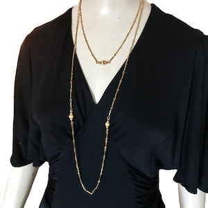 May include: A long gold chain necklace with two round beads. The necklace is layered and has a simple design.
