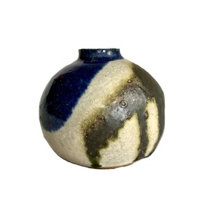 May include: A small, round ceramic vase with a narrow opening. The surface features a textured, off-white base glaze with prominent, irregular patches of deep navy blue and dark olive green that appear to drip down the sides of the vessel.
