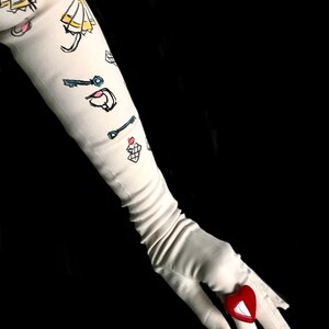 May include: A white long glove with a red heart-shaped ring on the finger. The glove has a pattern of keys, umbrellas, and other small drawings.