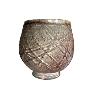 May include: A rounded ceramic cup with a textured, mottled brown and gray surface featuring a carved, intersecting geometric grid pattern. The vessel has a wide, open rim and a narrow, raised circular base, showcasing a rustic, earthy finish.