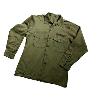 May include: An olive green long-sleeve button-down shirt featuring a pointed collar, two chest pockets with buttoned flaps, and the text U.S. ARMY embroidered in black thread above the right chest pocket. The shirt has buttoned cuffs.