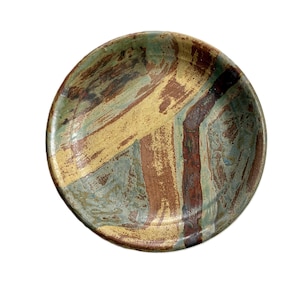 May include: A round ceramic plate with a hand-painted design. The plate features a mix of colors, including brown, yellow, and blue-green, with a textured, brushstroke pattern. The rim is a darker shade of brown.