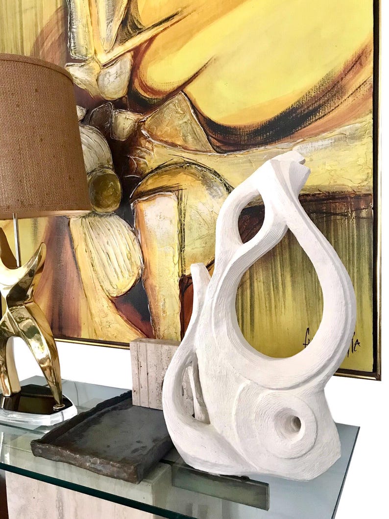 Pode incluir: Uma escultura abstrata branca com uma forma org&acirc;nica texturizada, apresentando uma grande abertura central e reentr&acirc;ncias circulares. A escultura tem um acabamento fosco e est&aacute; posicionada ao lado de uma bandeja retangular escura.