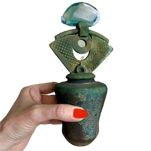 May include: A decorative bottle stopper featuring a textured, dark green and bronze-toned metallic base. The top section has a geometric, fan-like shape with a cross-hatched pattern and a central circular cutout, topped with a smooth, translucent blue-tinted agate stone.