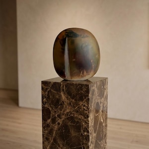 May include: A smooth, rounded, oval-shaped sculpture with a dark, mottled surface featuring blended tones of brown, green, and black. The sculpture rests atop a tall, rectangular pedestal made of polished brown marble with white veining.