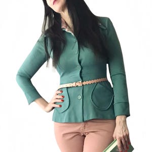 May include: A teal button-down jacket with a scalloped belt and peach-colored pants. The jacket has large, rounded pockets. A small book with the title "AMALFI" is held in the hand.