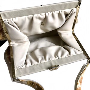 May include: A white clutch bag with a gold clasp and a gold chain strap. The bag has a white satin lining and the initials "JR" are printed on the inside.
