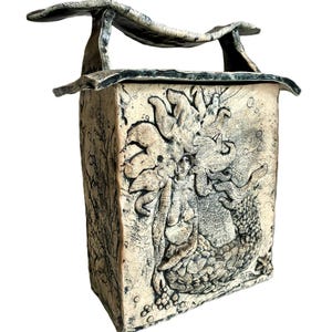 May include: A rectangular ceramic box with a textured, off-white finish and dark detailing. The front features a relief sculpture of a mermaid with flowing hair and a scaled tail. The lid has a curved, wave-like handle extending across the top.