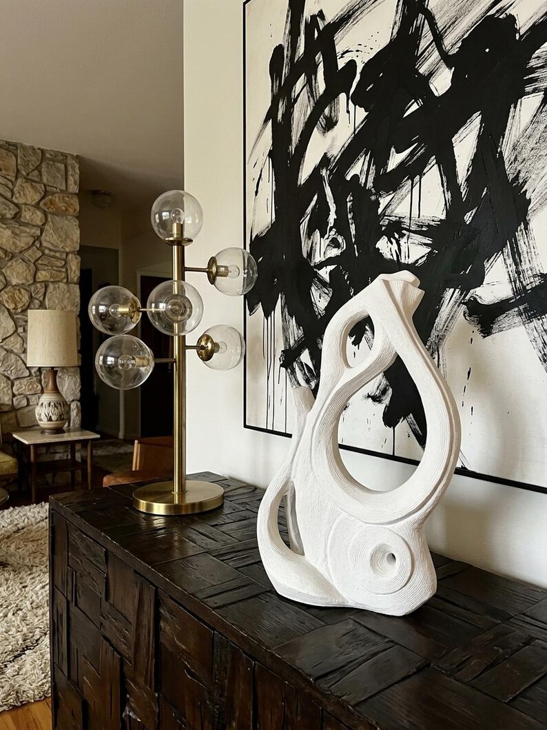 Pode incluir: Uma escultura abstrata branca com uma superf&iacute;cie texturizada e em camadas e recortes org&acirc;nicos curvos, posicionada ao lado de uma l&acirc;mpada de tom lat&atilde;o com v&aacute;rias l&acirc;mpadas de globo de vidro transparente sobre uma superf&iacute;cie de madeira escura.