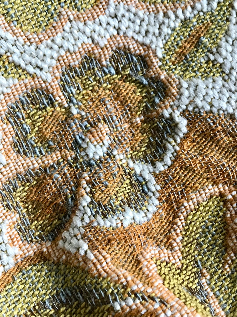 May include: Close-up of a woven fabric with a textured pattern in shades of yellow, orange, white, and grey.