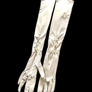 May include: A pair of long white satin gloves with pearl buttons. The gloves are fully extended and the fingers are spread out.